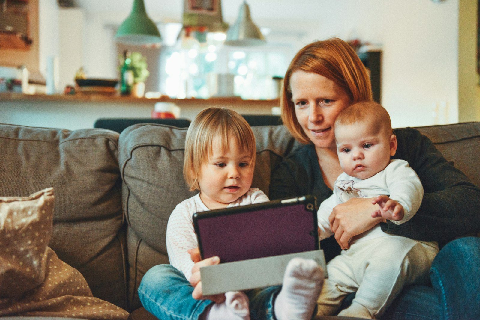 A mother on the couch with a toddler and a baby, the three of them looking at a tablet together