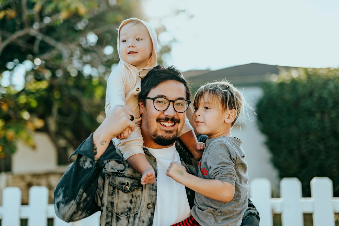 A father outside a house with a toddler on his shoulders and a small child in his arms